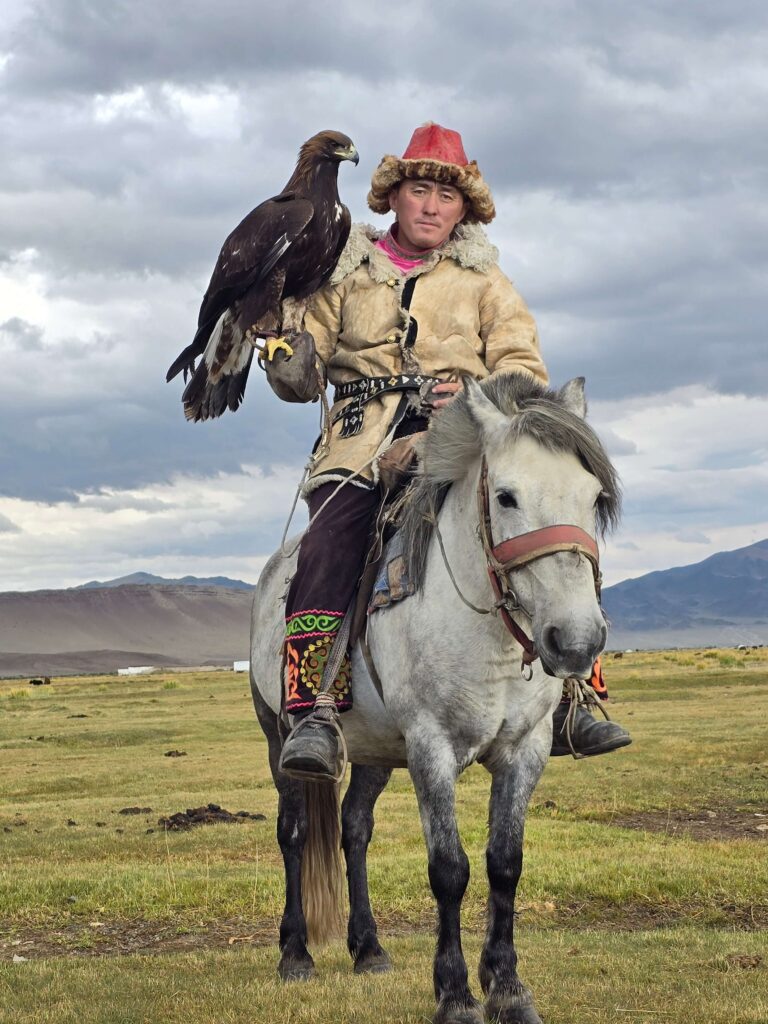 Kazakh eagle hunter in traditional dress on white horse holding golden eagle