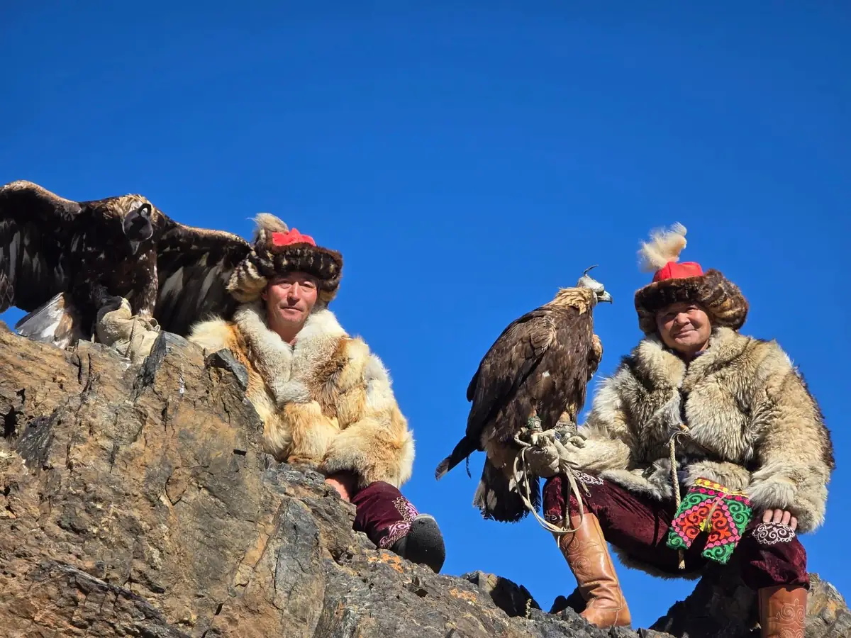 Two Kazakh eagle hunters in fur coats holding golden eagles on a rocky ridge in the Altai Mountains