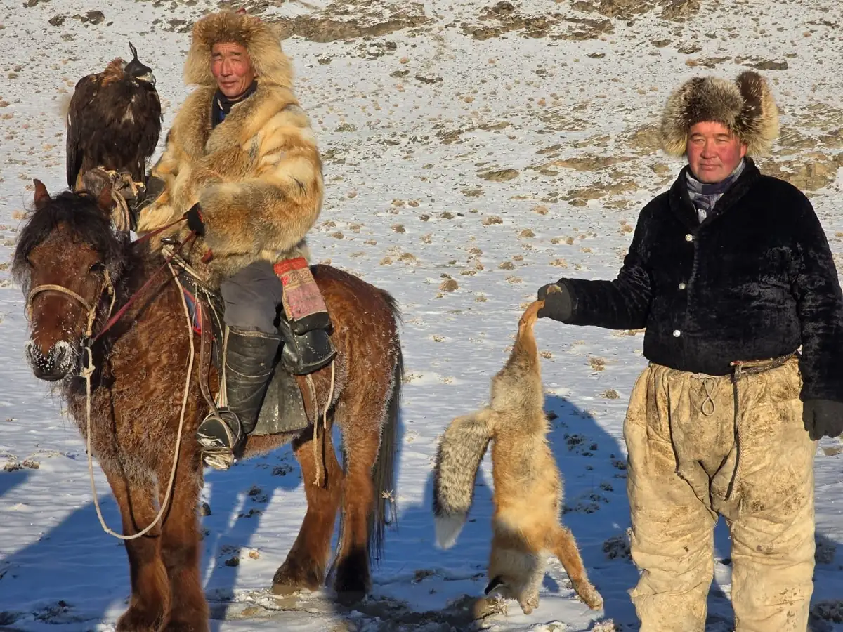 Two eagle hunters in fox-fur hats with a fox pelt on a snowy slope